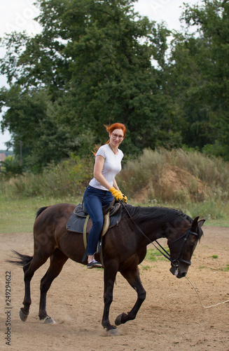 A redhead woman training riding a horse on the field. A green bush on the background
