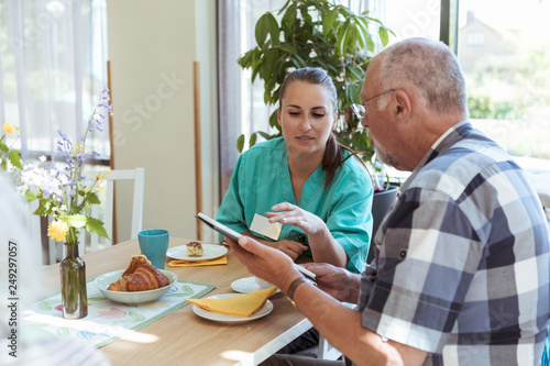 Young nurse and senior man using digital tablet at breakfast table in nursing home