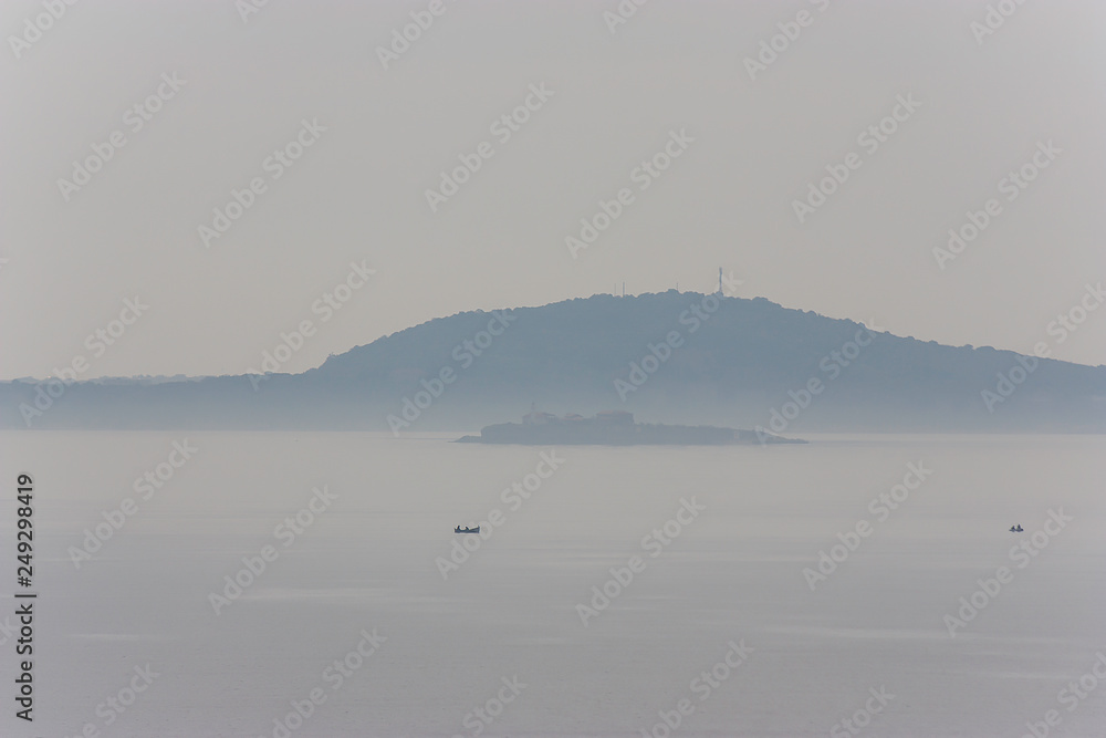 the island of Saint Anastasia in the Black Sea encircled with morning mist, and fishermen fish. Marine landscape in foggy weather early in the morning, before the blue hour.