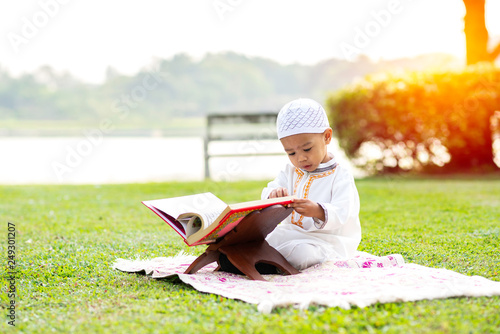 Little Muslim boy reading holy Quran on grass field near beautiful lake.
