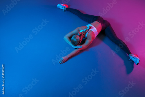 Attractive young slim gymnast woman in sports clothing stretching on the floor in neon lights. Flexible muscular woman doing gymnastic and yoga split.