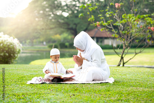 Muslim mother teaching her little son to reading the Quran on grass field near beautiful lake. Muslim family concept. 