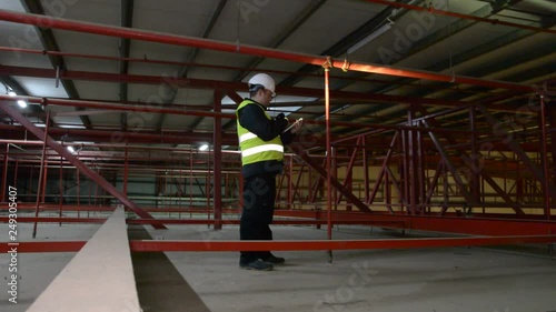 Technician engineer in a helmet checks the sprinklers pipes and roof constructions with a tablet