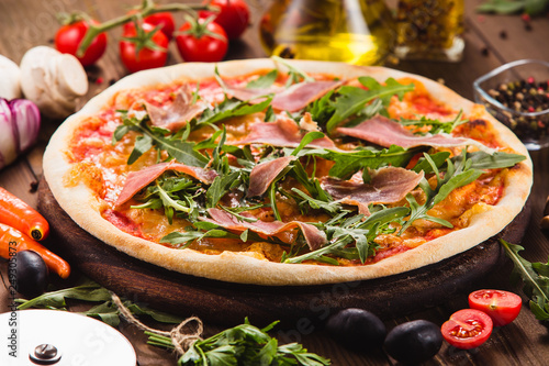 Pizza with bacon and arugula on a dark wooden background with ingredients around (close).