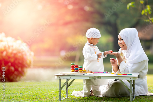 uslim mother teaching her little son to play math computation beads in the garden on grass field near beautiful lake. Muslim family concept.