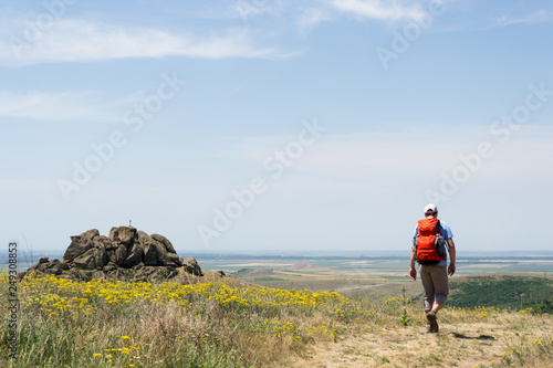 Hiker with a red backpack on a trail with yellow flowers  in Macin Mountains  oldest mountains in Romania  passing near rocks with a small cross on top of them  during a sunny day with clear skies.