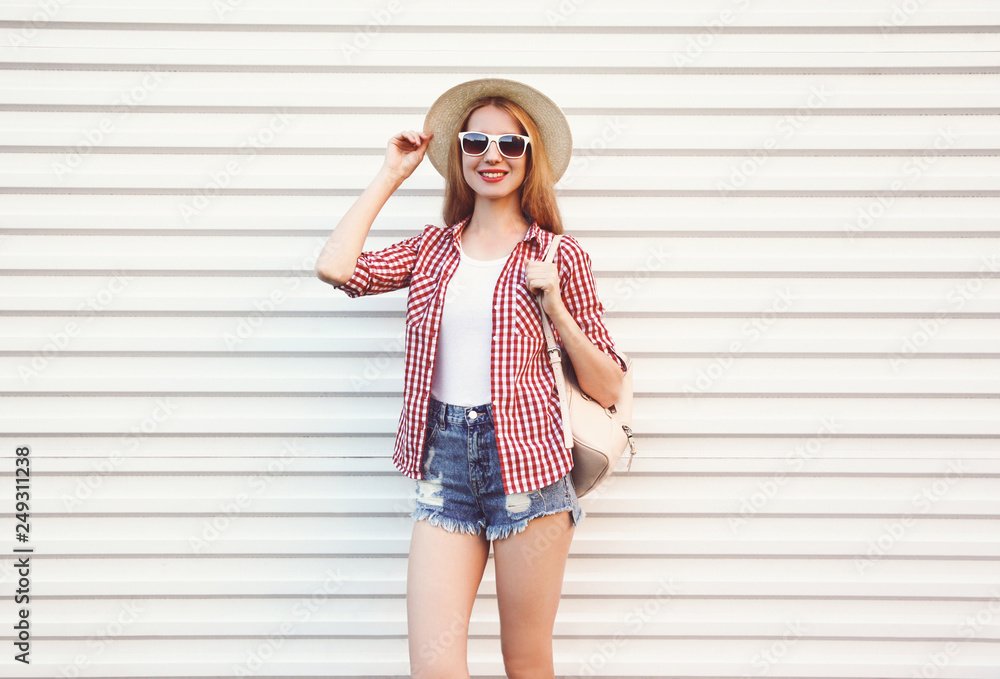 Happy smiling young woman in summer round straw hat, checkered shirt, shorts posing on white wall background