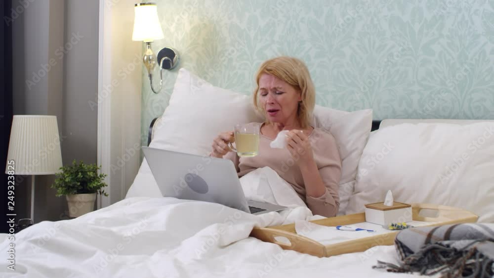Medium shot of coughing woman resting in bed and drinking hot water with lemon while talking to online doctor using telemedicine service on laptop Medium shot of coughing woman resting in bed and drinking hot water with lemon while talking to online doctor using telemedicine service on laptop