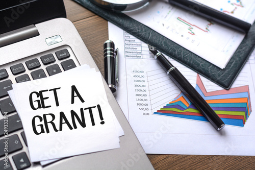 Desk with business accessories, calculator, documents with charts, fountain pen, laptop and paper for notes with text Get a grant