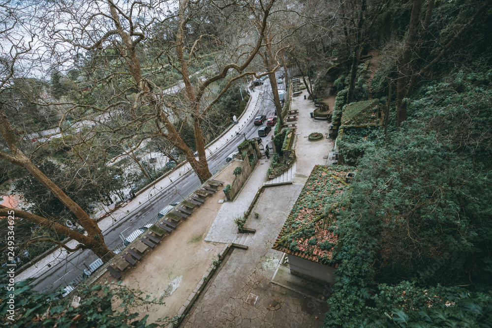 Fototapeta premium A view from above of the park in Sintra, Portugal, near a bending road with cars: a cracked stony floor, beautiful trees, antique tiled roofs of outbuildings, row of tables with benches; wide-angle