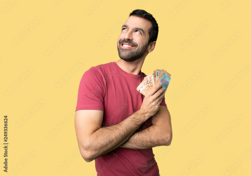 Man taking a lot of money looking up while smiling on isolated yellow background Man taking a lot of money looking up while smiling on isolated yellow background