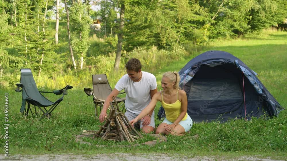 Smiling young Caucasian woman and man high five after completing a campfire by their tent. Boyfriend and girlfriend gathering firewood and prepare a campfire. Cheerful travelers camping in calm nature Smiling young Caucasian woman and man high five after completing a campfire by their tent. Boyfriend and girlfriend gathering firewood and prepare a campfire. Cheerful travelers camping in calm nature