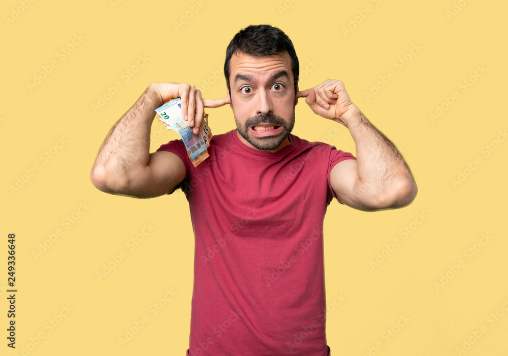 Man taking a lot of money covering both ears with hands on isolated yellow background Man taking a lot of money covering both ears with hands on isolated yellow background