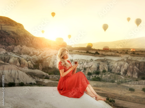 A girl sitting on the top of a cliff with a glass of Turkish tea at dawn with a view of the mountains of Cappadocia and balloons in the sky. The spirit of travel and happy holidays.