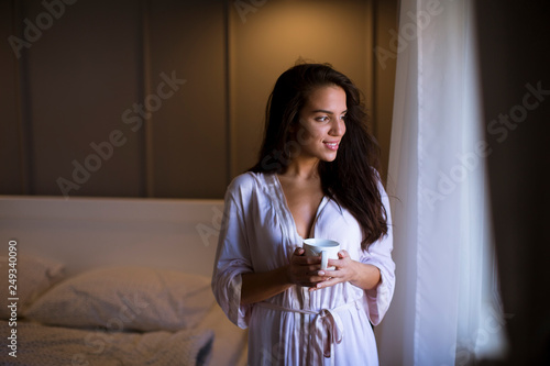Young woman with cup of coffee or tea in neglige standing by the window