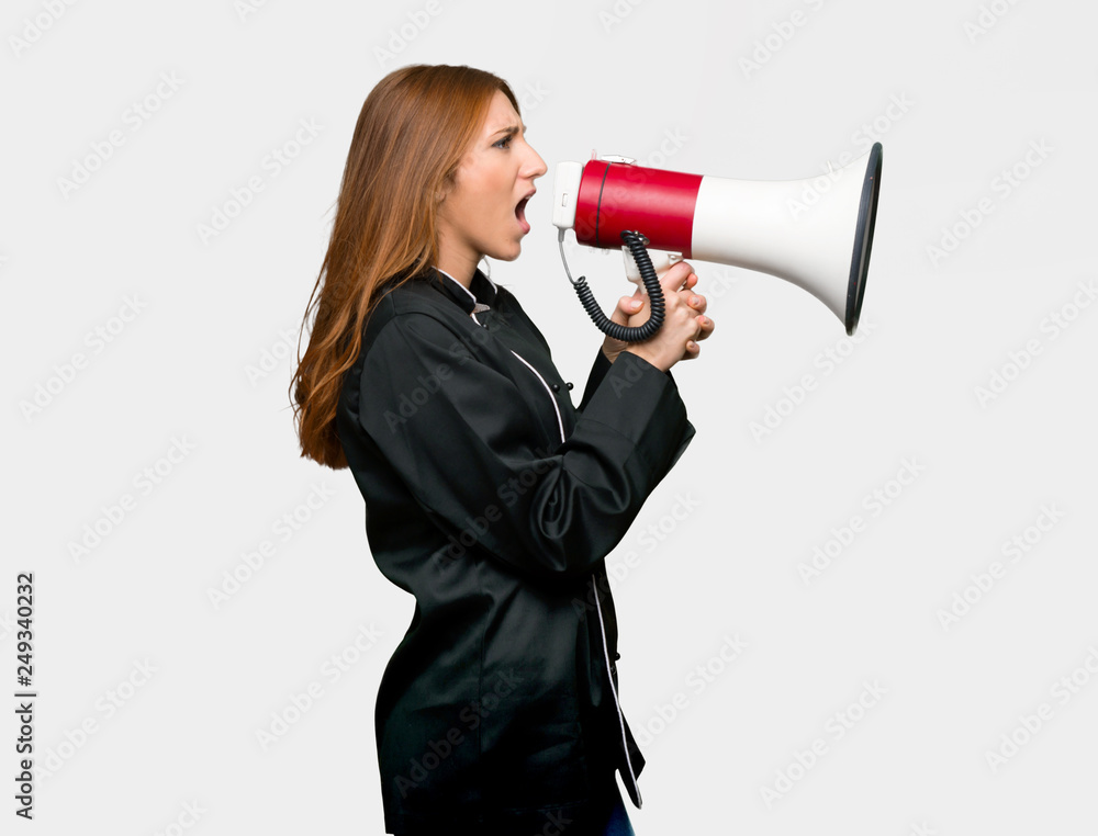 Young redhead chef woman shouting through a megaphone to announce something in lateral position on isolated grey background Young redhead chef woman shouting through a megaphone to announce something in lateral position on isolated grey background