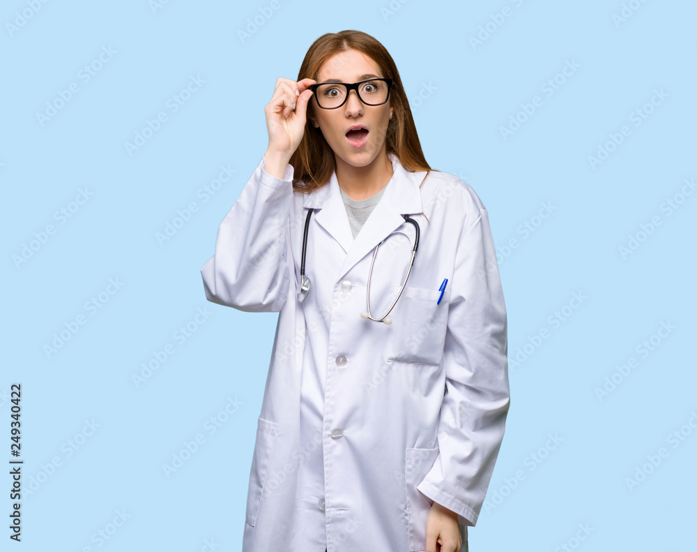 Redhead doctor woman with glasses and surprised on isolated blue background Redhead doctor woman with glasses and surprised on isolated blue background