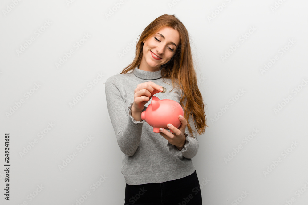 Redhead girl over white wall taking a piggy bank and happy because it is full Redhead girl over white wall taking a piggy bank and happy because it is full