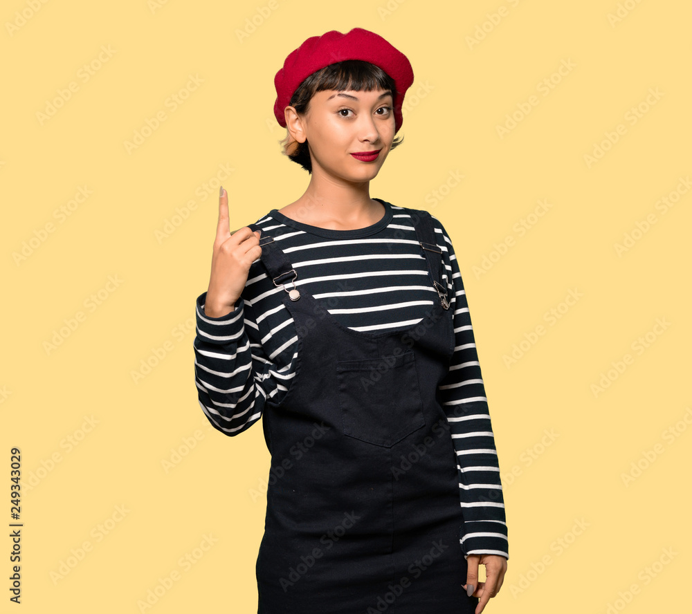 Young woman with beret showing and lifting a finger in sign of the best over yellow background Young woman with beret showing and lifting a finger in sign of the best over yellow background