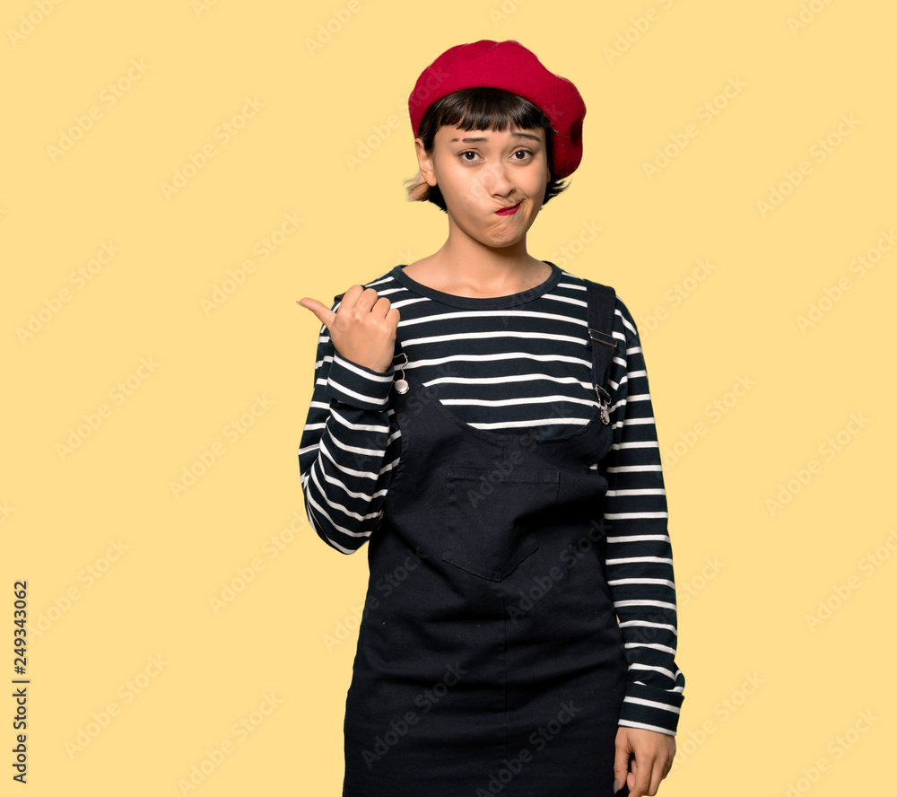 Young woman with beret unhappy and pointing to the side over yellow background