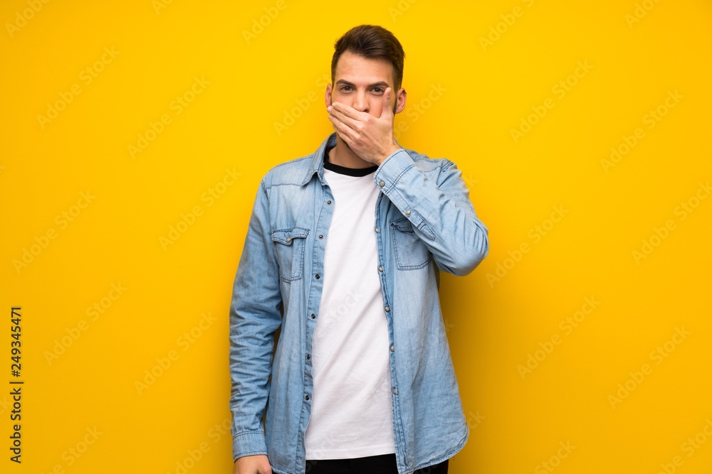Handsome man over yellow wall covering mouth with hands for saying something inappropriate Handsome man over yellow wall covering mouth with hands for saying something inappropriate
