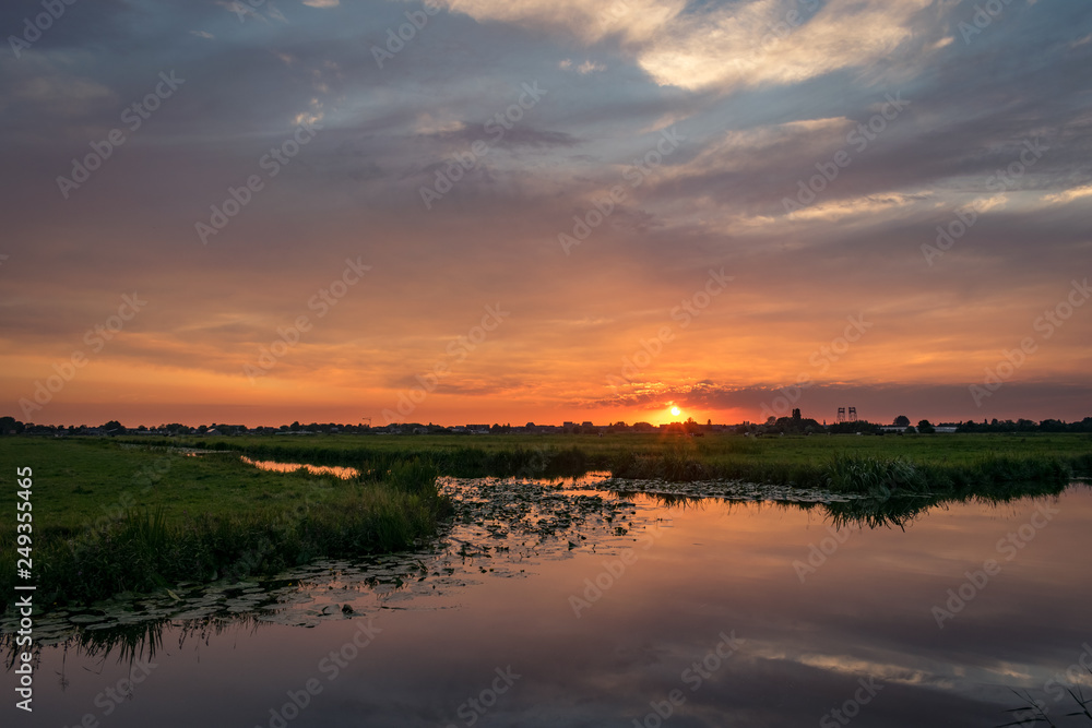 The sun sets on the horizon of the dutch countryside. A beautiful colored sky is reflected in the water of a lake. 