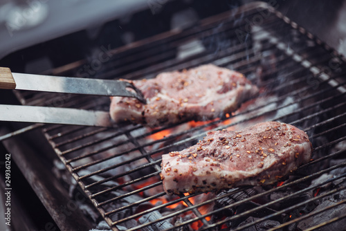 selective focus of juicy raw steaks grilling on barbecue grid with smoke and tweezers