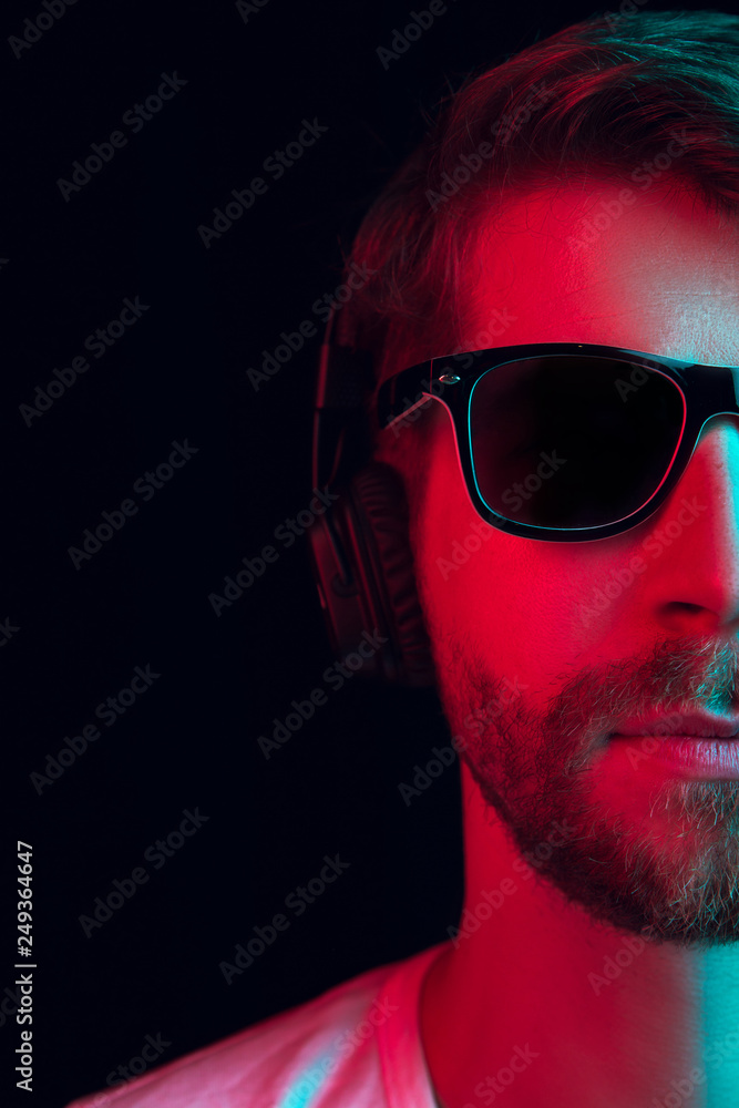 Enjoying his favorite music. Happy young stylish man in sunglasses with headphones listening sound and smiling while standing against blue neon background Enjoying his favorite music. Happy young stylish man in sunglasses with headphones listening sound and smiling while standing against blue neon background