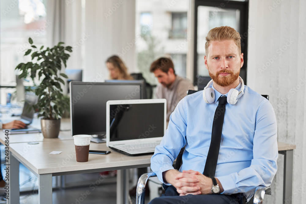 Serious CEO in formal wear and with headphones around neck sitting in chair and looking at camera. Office interior.