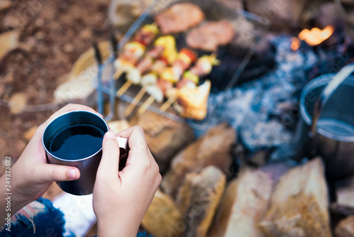 Close up woman hand with a coffee cup with grilling barbecue background in the campground at summer camp travel, Skewers of pork in camping, Summer Camp Travel one activity for relaxing.