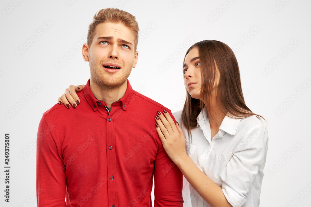 a pretty brunette in a white shirt hugs and reassures her companion - a young man with blond hair who rolls eyes up in horror and fatigue. Studio portrait on isolated white background. a pretty brunette in a white shirt hugs and reassures her companion - a young man with blond hair who rolls eyes up in horror and fatigue. Studio portrait on isolated white background.