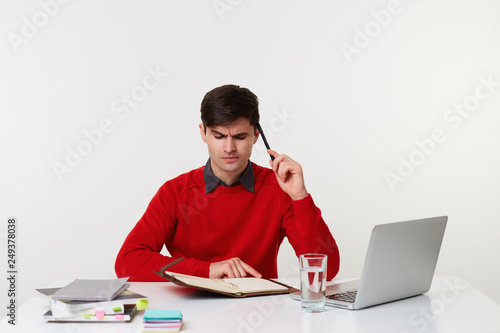 Brunet man intensely looks at his notes in the diary, makes up a schedule, looks thoughtfully into his notebook, sits at the desk on which the laptop books lay, is isolated on a white background