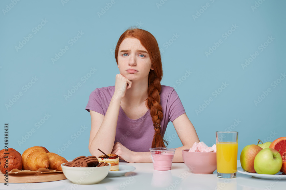 Sad upset red-haired girl looking camera with dissatisfaction, thinks about diet, extra calories, baking food and fresh fruit juice yogurt lay on the table, on a blue background. Healthy food concept Sad upset red-haired girl looking camera with dissatisfaction, thinks about diet, extra calories, baking food and fresh fruit juice yogurt lay on the table, on a blue background. Healthy food concept