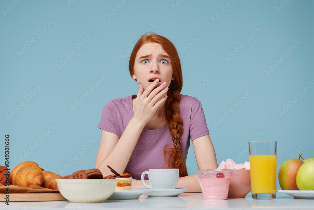 Frightened scared red-haired girl covers open mouth with a palm, sits at a table on which there are many different foods baking fresh fruit,worrying about her health figure, diet violation Frightened scared red-haired girl covers open mouth with a palm, sits at a table on which there are many different foods baking fresh fruit,worrying about her health figure, diet violation