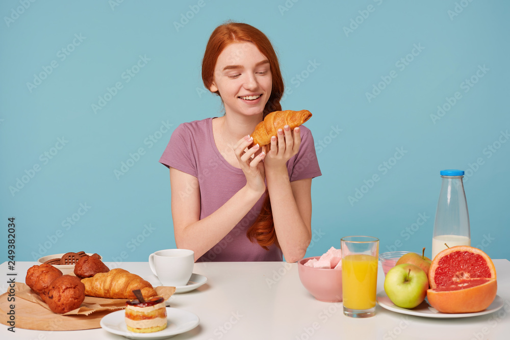 Cute red-haired girl with a braid sits at a table, has breakfast, with excitement pleasure looks at croissant,pastries on the table and fresh fruit,on a blue background Cute red-haired girl with a braid sits at a table, has breakfast, with excitement pleasure looks at croissant,pastries on the table and fresh fruit,on a blue background