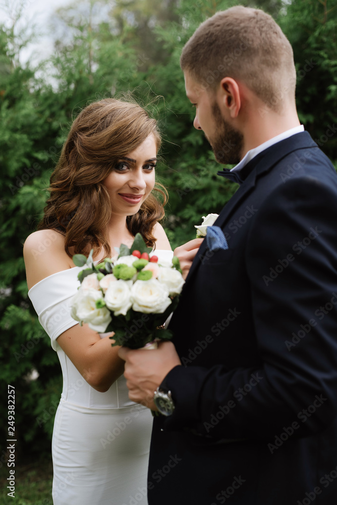 Two young beautiful lovers getting ready for the wedding outdoor on green background. Pretty bride putting on the buttonhole bouquet. wedding concept Two young beautiful lovers getting ready for the wedding outdoor on green background. Pretty bride putting on the buttonhole bouquet. wedding concept