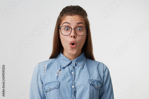 Close up face of a young girl with long hair in glasses looks surprised, amazed, shoked. Woman found out incredible news, witnessed unforgettable event on a white background photo