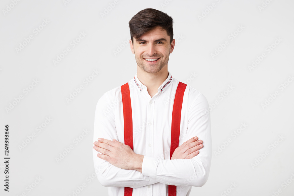 Close up young businessman with dark hair unshaven standing with his arms crossed, wearing a white shirt, red suspenders, smiling looks confident glad and happy isolated on a white background. Close up young businessman with dark hair unshaven standing with his arms crossed, wearing a white shirt, red suspenders, smiling looks confident glad and happy isolated on a white background.