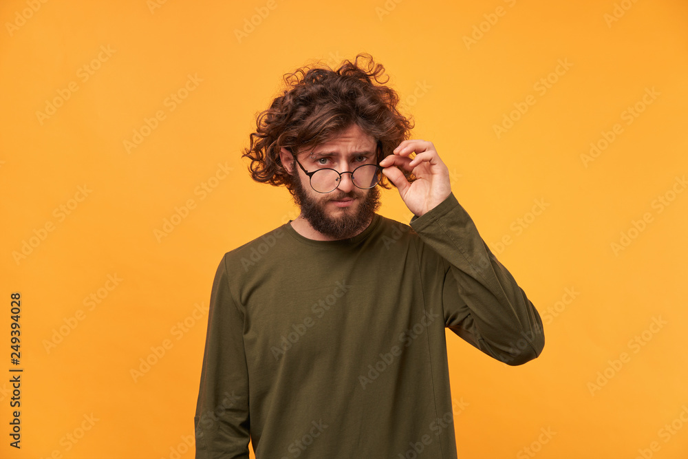 Close up of young man with a beard and curly dark hair, with interest lowered his glasses, carefully considering something, isolated on a yellow background Close up of young man with a beard and curly dark hair, with interest lowered his glasses, carefully considering something, isolated on a yellow background