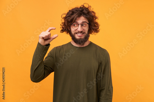 Handsome unshaven male with dark curly hair and thick bristle, shows something tiny with hands, dressed casually, isolated over yellow background. Young man demonstrates small thing indoor.
