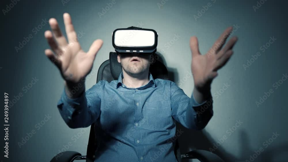 A man in a shirt sits in an armchair and watches a movie with virtual reality glasses. Surprised guy in VR headset waving his arms. A man in a shirt sits in an armchair and watches a movie with virtual reality glasses. Surprised guy in VR headset waving his arms.
