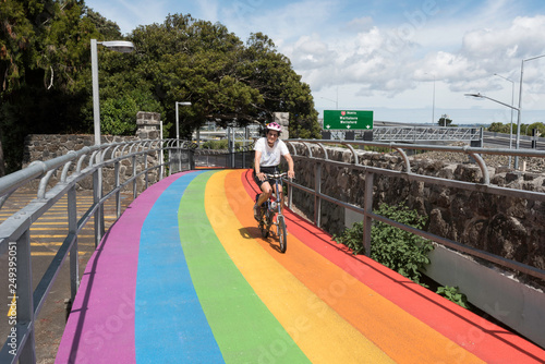 Female, baby boomer cyclist crossing a bridge on a bike path painted in rainbow colours in Waterview, Auckland, New Zealand.