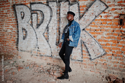 African american man in jeans jacket, beret and eyeglasses against graffiti brick wall with bruk sign.