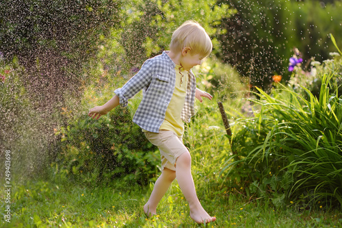 Funny little boy playing with garden sprinkler in sunny backyard