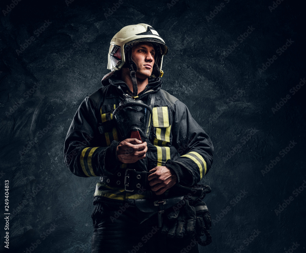 Studio portrait in a dark studio against a textured wall. Brutal firefighter in uniform and safety helmet holding an oxygen mask and looking sideways with a confident look. Studio portrait in a dark studio against a textured wall. Brutal firefighter in uniform and safety helmet holding an oxygen mask and looking sideways with a confident look.