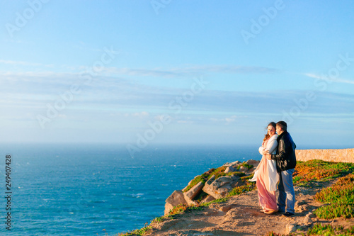 A man hugs a woman from the back side on the rocky shore of the ocean