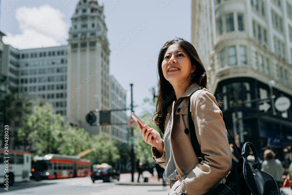 Fototapeta premium fashion happy woman tourist standing under blue sky in busy city area in san francisco. young girl smiling looking aside holding cellphone online map on city street on sunny day. charming lady laugh