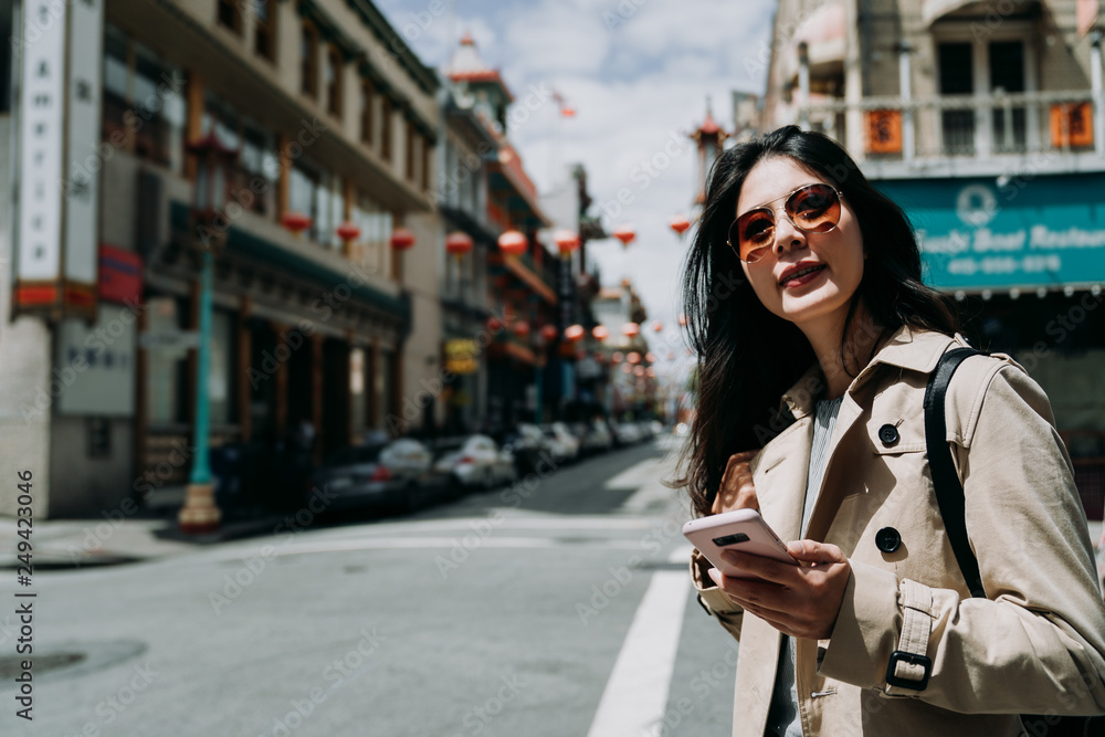 Happy young woman in sunglasses enjoying weekends spending time outdoors holding smartphone for chatting. cheerful girl laughing on street looking away mobile phone in hand walking in chinatown.