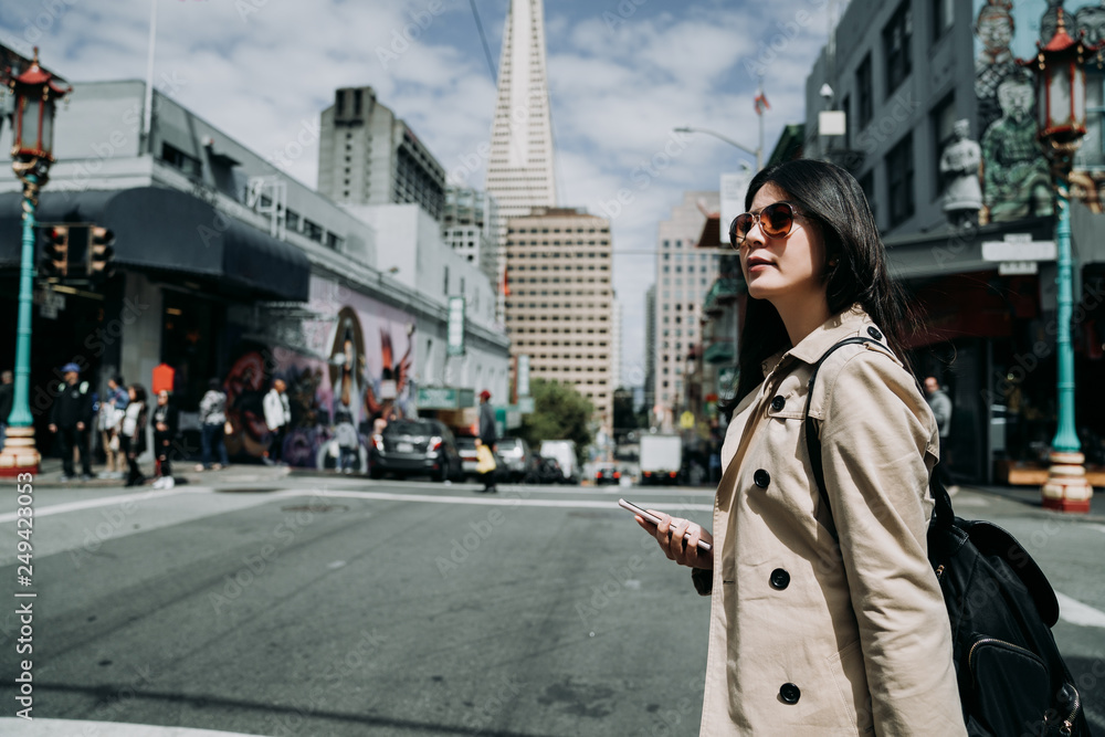 pretty cool smiling young asian woman using smartphone walking in city san francisco. happy female model wearing elegant sunglasses outdoors sightseeing in chinatown. Transamerica Pyramid in back.
