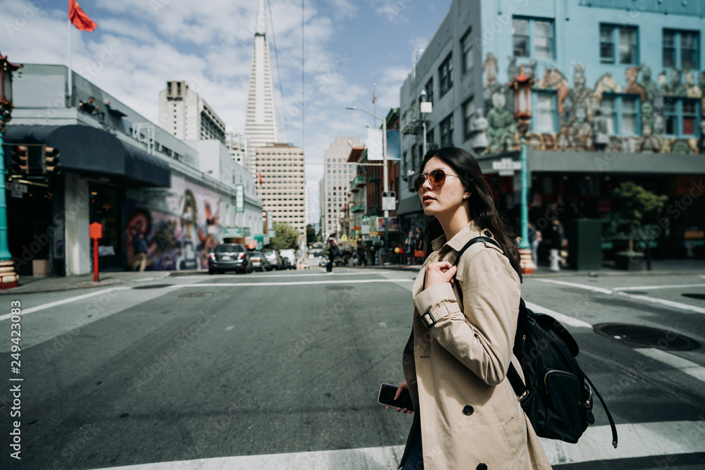 Fototapeta premium Beautiful asian travel woman wearing fashionable sunglasses holding smart phone in hand. young girl backpacker walking on street in chinatown on sunny day. tall tower Transamerica Pyramid in back.